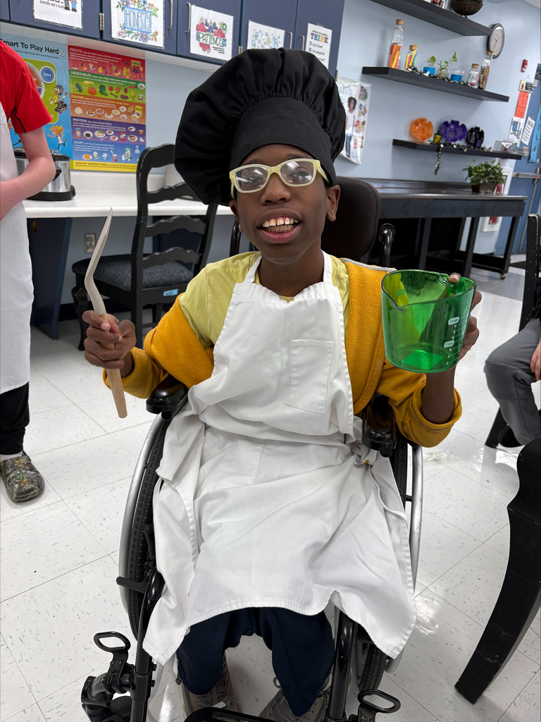 A student wearing a chef hat and apron holds cooking tools while participating in a hands-on classroom cooking activity focused on life skills and food preparation.