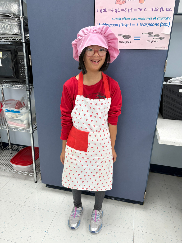 A student stands in a classroom kitchen wearing a pink chef hat, glasses, and a heart-patterned apron. The student is dressed for a hands-on cooking lesson and stands in front of a cabinet. A poster about measuring capacity is displayed on the wall nearby, supporting instruction in cooking and life skills.
