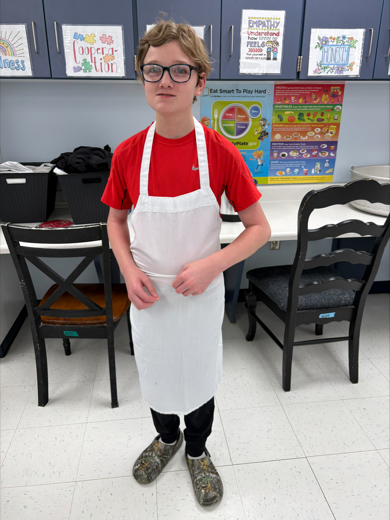 A student wearing glasses and a white apron stands in a classroom kitchen prepared for a cooking activity. Educational posters about nutrition and character traits are displayed on the wall behind the student. The hands-on lesson supports the development of cooking, life, and workplace readiness skills in an inclusive learning environment