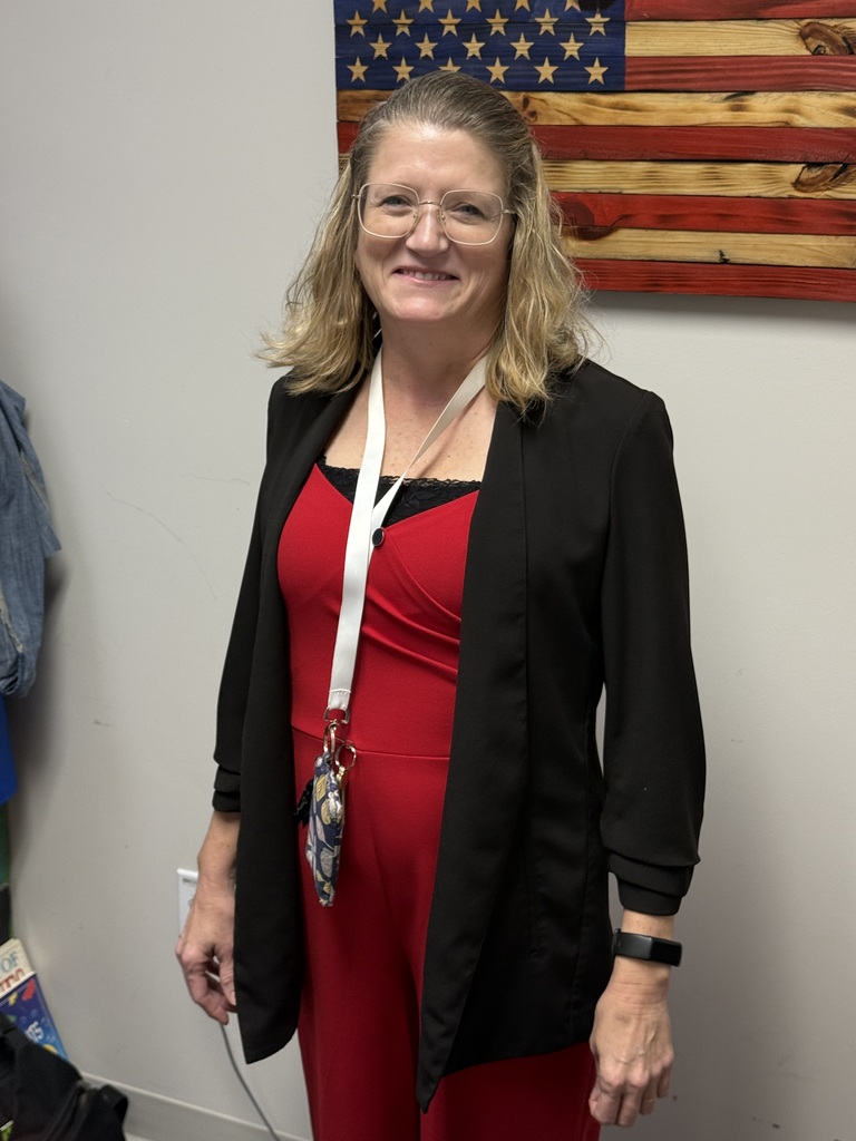 A staff member wearing a red dress and black cardigan stands indoors in an office or conference room during Red Ribbon Spirit Day. A lanyard with identification hangs around the staff member's neck. A decorative wooden American flag is mounted on the wall behind them. The staff member is dressed in red to support the school's commitment to promoting healthy, drug-free choices and school spirit.