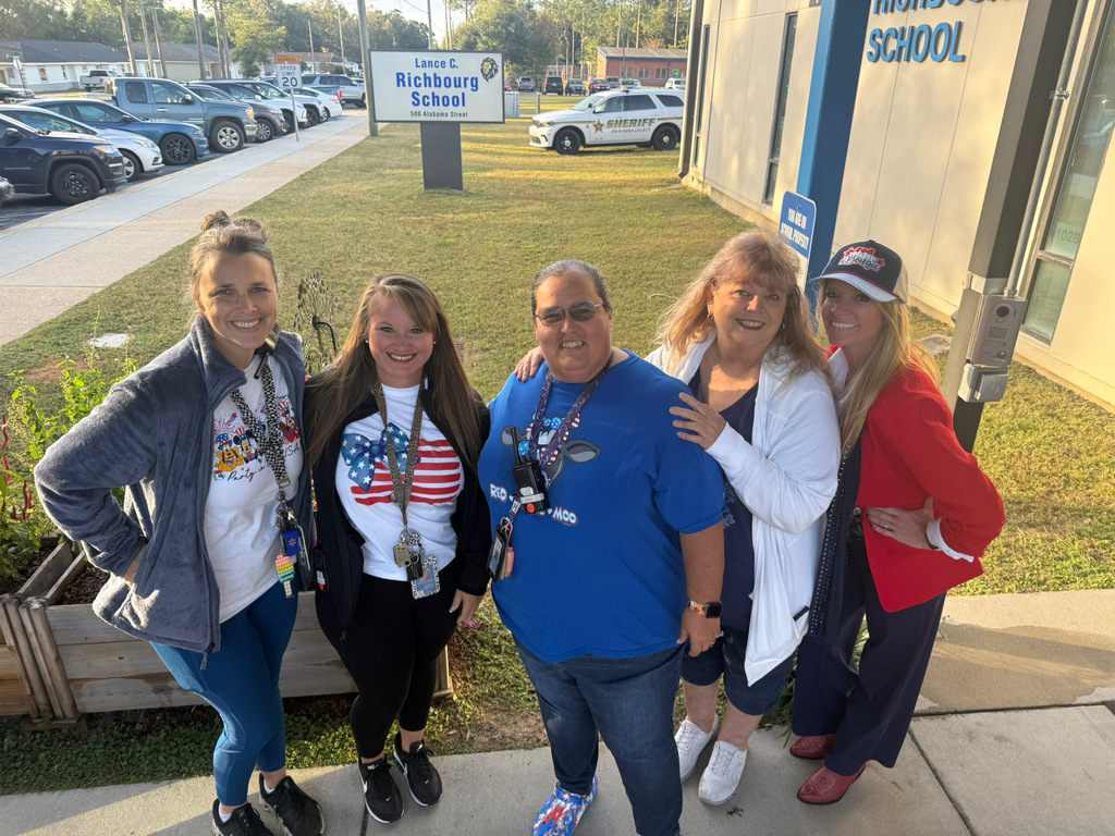 Five smiling Richbourg School staff members pose together outside the school near the Lance C. Richbourg School sign. The group wears a variety of red, white, and blue clothing, including patriotic shirts, blue jeans, and red accessories, in celebration of Red Ribbon Week's “Stand Proud to Be Drug Free” spirit day.