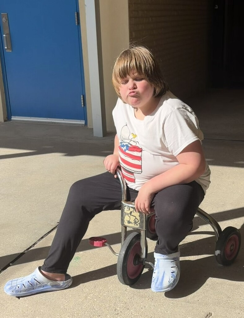 A Richbourg student with short, light brown hair sits outside in the sunshine on an adaptive tricycle, wearing a white T-shirt with a red, white, and blue patriotic design, black pants, and light blue shoes. The student makes a playful face while posing for a photo during Red Ribbon Week's “Stand Proud to Be Drug Free” spirit day.