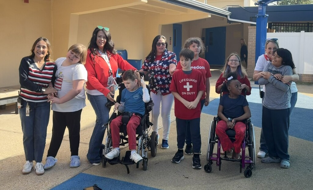 A group of Richbourg students and staff pose together outside the school in celebration of Red Ribbon Week's “Stand Proud to Be Drug Free” spirit day. Several participants wear patriotic red, white, and blue clothing, including stars, stripes, and red shirts. Two students are seated in wheelchairs, while others stand alongside staff members. The group gathers in a sunny outdoor courtyard to show their school spirit.