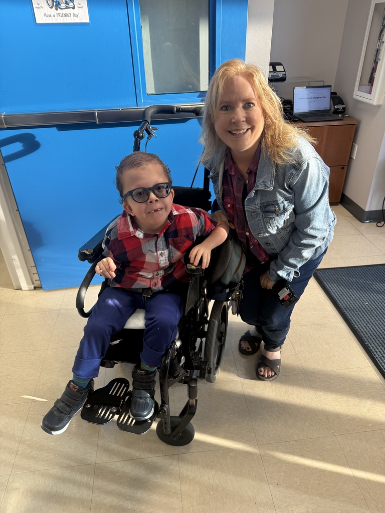A smiling young student wearing glasses, a red, white, and blue plaid shirt, and blue pants sits in a wheelchair beside a smiling staff member wearing a denim jacket over a red plaid shirt. They pose together in a school hallway near bright blue doors.