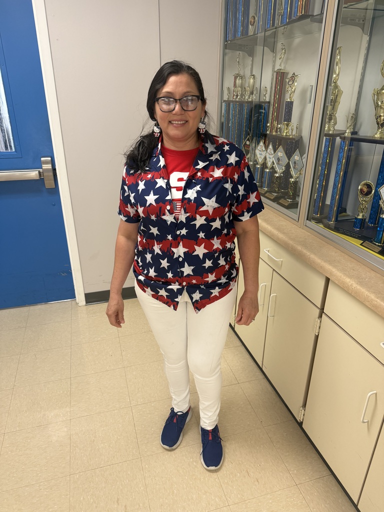 A smiling Richbourg staff member wearing glasses, a red, white, and blue star-patterned shirt, white pants, and navy blue shoes poses in the school hallway beside a glass trophy display case. Her patriotic outfit celebrates Red Ribbon Week's “Stand Proud to Be Drug Free” spirit day.