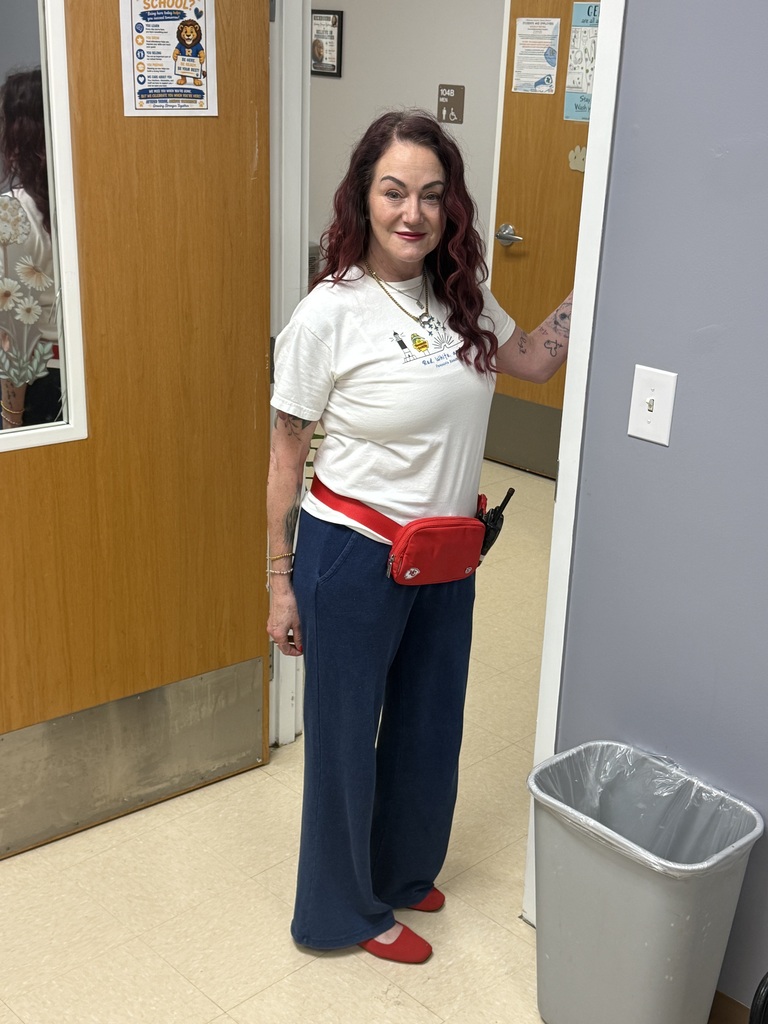 A smiling Richbourg staff member with long, dark red hair stands in a school doorway wearing a white T-shirt, navy blue pants, red shoes, and a red waist bag. Her red, white, and blue outfit shows school spirit in celebration of Red Ribbon Week's “Stand Proud to Be Drug Free” spirit day.
