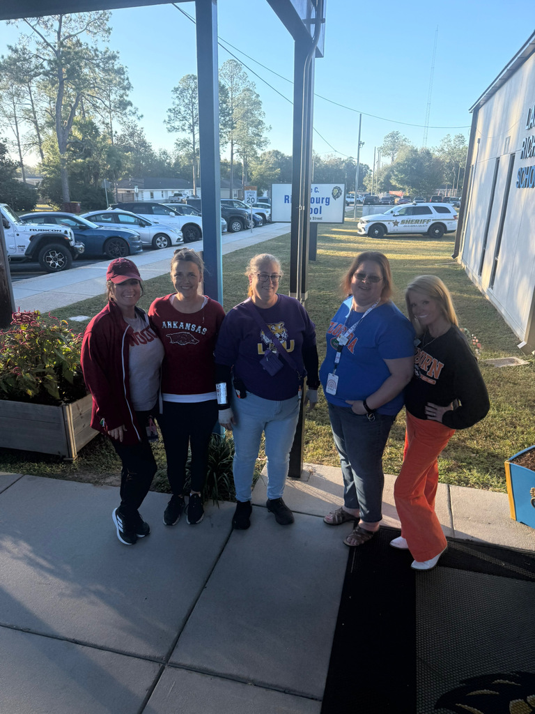 Five smiling Richbourg School staff members pose together outside the school wearing their favorite college and school team shirts, including Arkansas, LSU, Florida, and Auburn. The group stands near the school entrance with the Richbourg School sign visible in the background, celebrating Red Ribbon Week's “Team Up Against Drugs” spirit day.