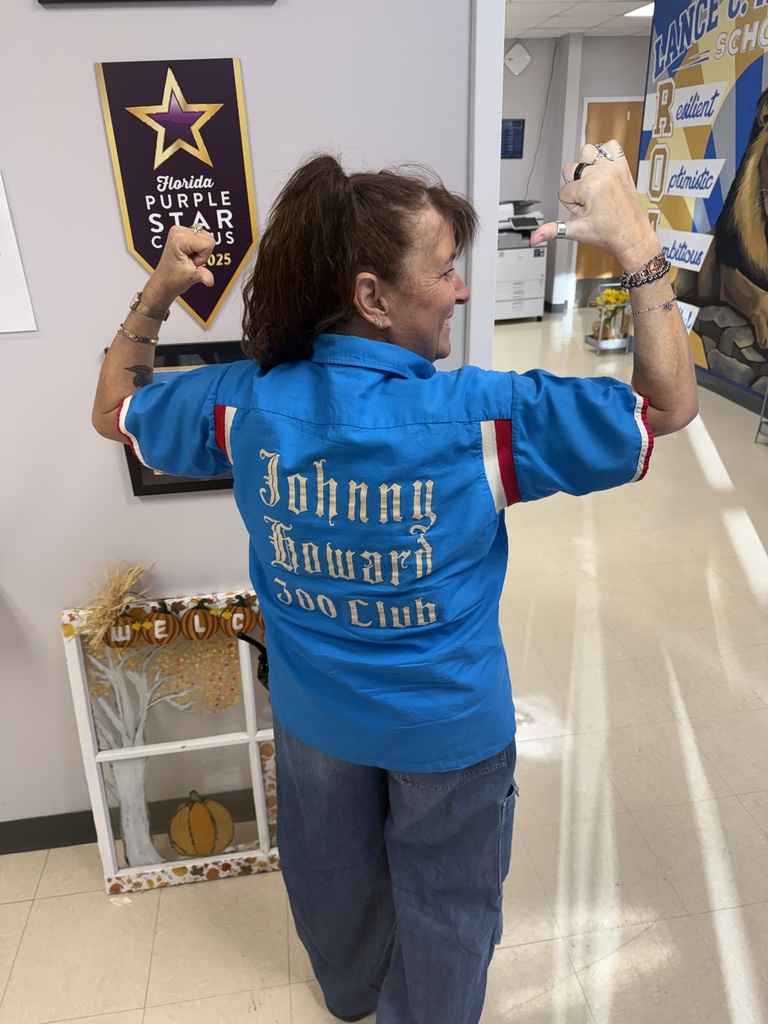 A smiling Richbourg School staff member poses in the school hallway with her back toward the camera, raising both arms to show off her grandfather's bright blue “Johnny Howard Zoo Club” bowling jersey. She stands beside a Florida Purple Star Campus banner and a fall decoration while celebrating Red Ribbon Week's “Team Up Against Drugs” spirit day.