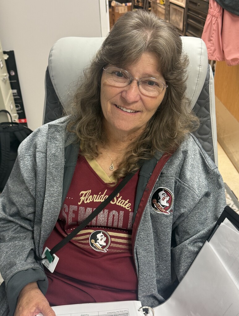 A smiling Richbourg School staff member with glasses and shoulder-length brown hair sits in an office chair wearing a garnet Florida State Seminoles shirt and a gray Florida State jacket. She holds an open binder while showing her team spirit during Red Ribbon Week's “Team Up Against Drugs” spirit day.