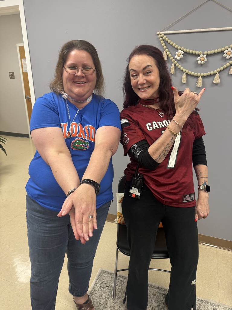 Two smiling Richbourg School staff members pose together indoors during Red Ribbon Week's “Team Up Against Drugs” spirit day. One wears a blue Florida Gators T-shirt and jeans, while the other wears a garnet South Carolina Gamecocks football jersey with the number 7 and black pants. Both proudly show their team spirit while posing for the photo.