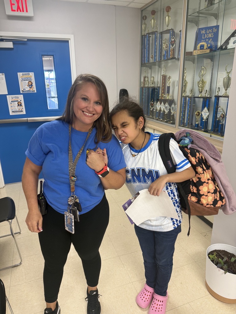 A smiling Richbourg School staff member wearing a blue shirt and black pants stands beside a student wearing a white and blue sports jersey, navy pants, and pink Crocs. The two hold hands and pose playfully in the school hallway near a trophy display case. The student carries a floral backpack and papers while celebrating Red Ribbon Week's “Team Up Against Drugs” spirit day.