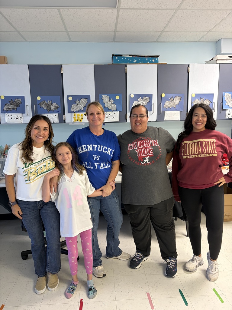 Four smiling Richbourg School staff members and one student pose together in a classroom during Red Ribbon Week's “Team Up Against Drugs” spirit day. The group wears shirts representing the University of South Florida Bulls, Kentucky Wildcats, Alabama Crimson Tide, and Florida State University. The student wears a white jersey with the number 14 and pink leggings. The group stands in front of classroom cabinets decorated with student artwork featuring bats.