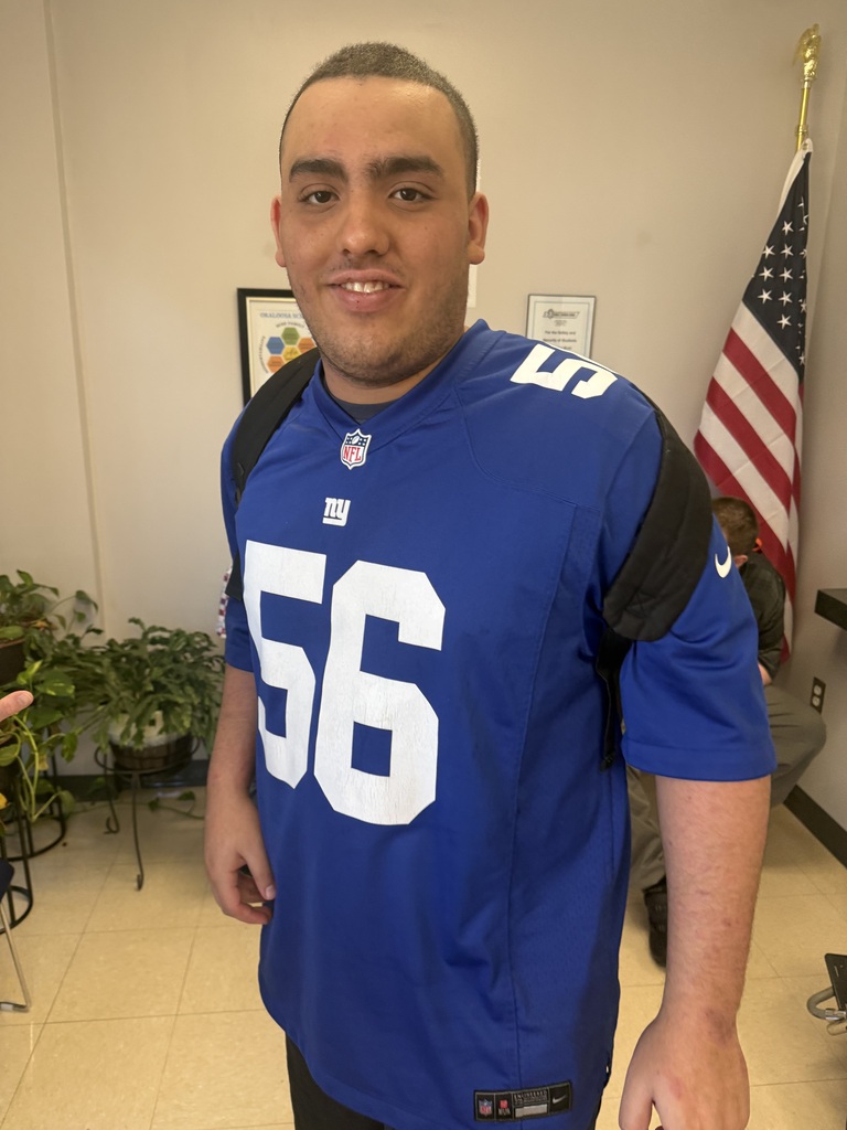 A smiling Richbourg School student poses indoors wearing a blue New York Giants football jersey with the number 56 and a black backpack. An American flag is visible in the background. The student proudly shows his team spirit during Red Ribbon Week's “Team Up Against Drugs” spirit day.