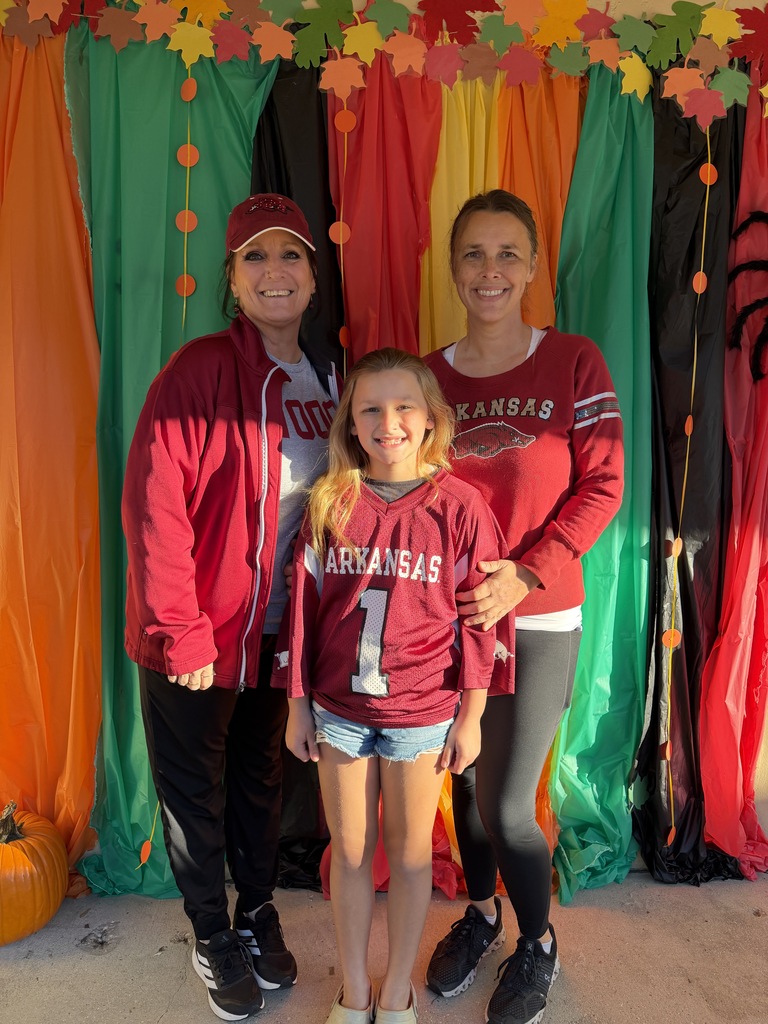Two smiling Richbourg School staff members pose with a smiling student in front of a colorful fall-themed backdrop decorated with autumn leaves and pumpkins. All three wear Arkansas Razorbacks apparel in cardinal red, with the student wearing a football jersey featuring the number 1. They proudly show their team spirit during Red Ribbon Week's “Team Up Against Drugs” spirit day.