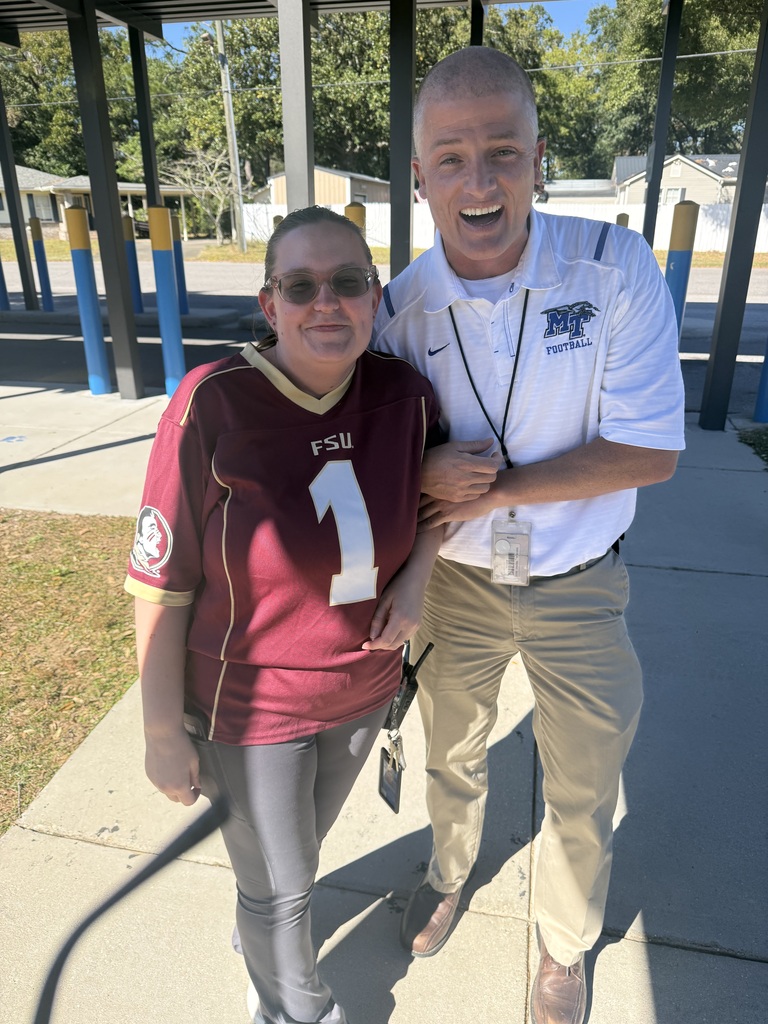 Two smiling Richbourg School staff members pose together outside under a covered walkway during Red Ribbon Week's “Team Up Against Drugs” spirit day. One wears a garnet Florida State University (FSU) football jersey with the number 1, while the other wears a white Middle Tennessee (MT) football polo shirt and khaki pants. Both proudly show their team spirit in support of making healthy, drug-free choices.