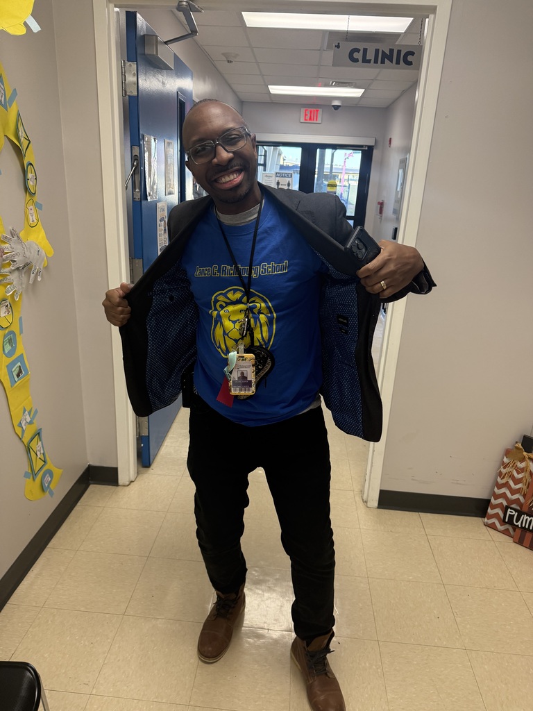 A smiling Richbourg School staff member wearing glasses stands in the school hallway near the clinic, holding open his black jacket to proudly display a royal blue Lance C. Richbourg School T-shirt featuring the school's yellow lion mascot. He celebrates Red Ribbon Week's “Team Up Against Drugs” spirit day by showing his Richbourg Lions school pride.