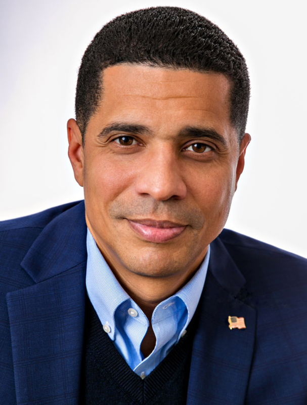 Superintendent Marcus Chambers'  professional  head-and-shoulders portrait against a plain white background, featuring a person wearing a dark blue blazer layered over a dark sweater vest and a light blue collared shirt, with a small U.S. flag pin attached to the blazer lapel.