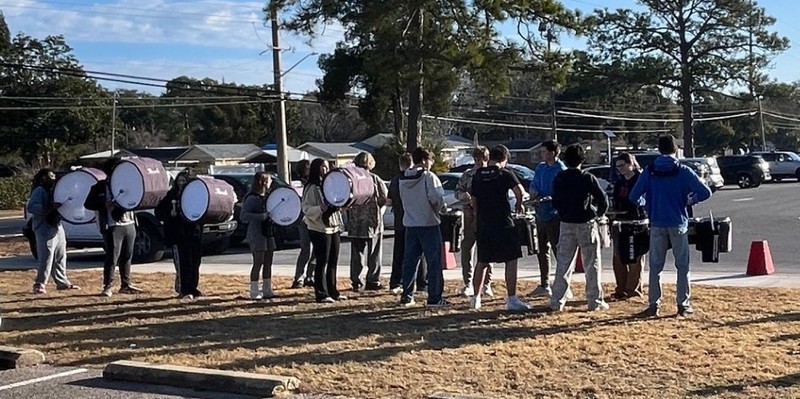 A group of students stands on a grassy area near a parking lot, holding and playing marching band drums. Cars and trees are visible in the background on a sunny day.