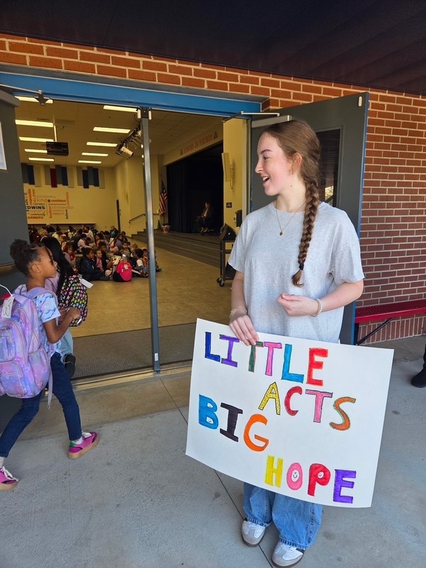 A person stands outside the open doors of a school auditorium, holding a brightly colored sign that reads “LITTLE ACTS BIG HOPE.” Inside the auditorium, many children sit on the floor, facing the stage. A few students with backpacks walk past the doorway.