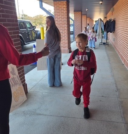 A young child in a red outfit walks along a covered sidewalk outside a brick school building, carrying a backpack. Adults stand nearby holding signs as students enter the school. Coats and jackets hang on a rack against the wall.