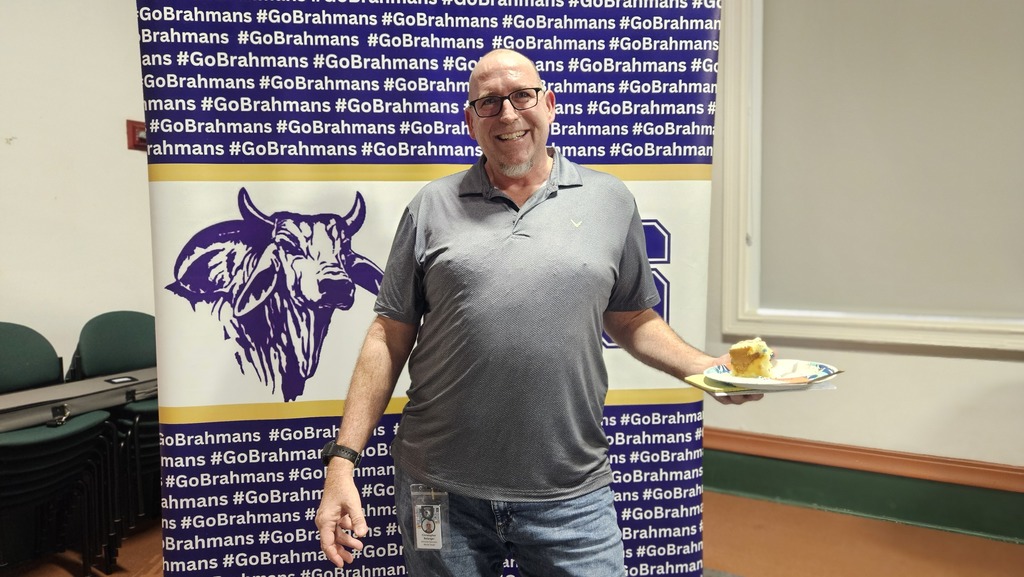 one teacher wearing gray shirt and holding cake Infront of brahman backdrop