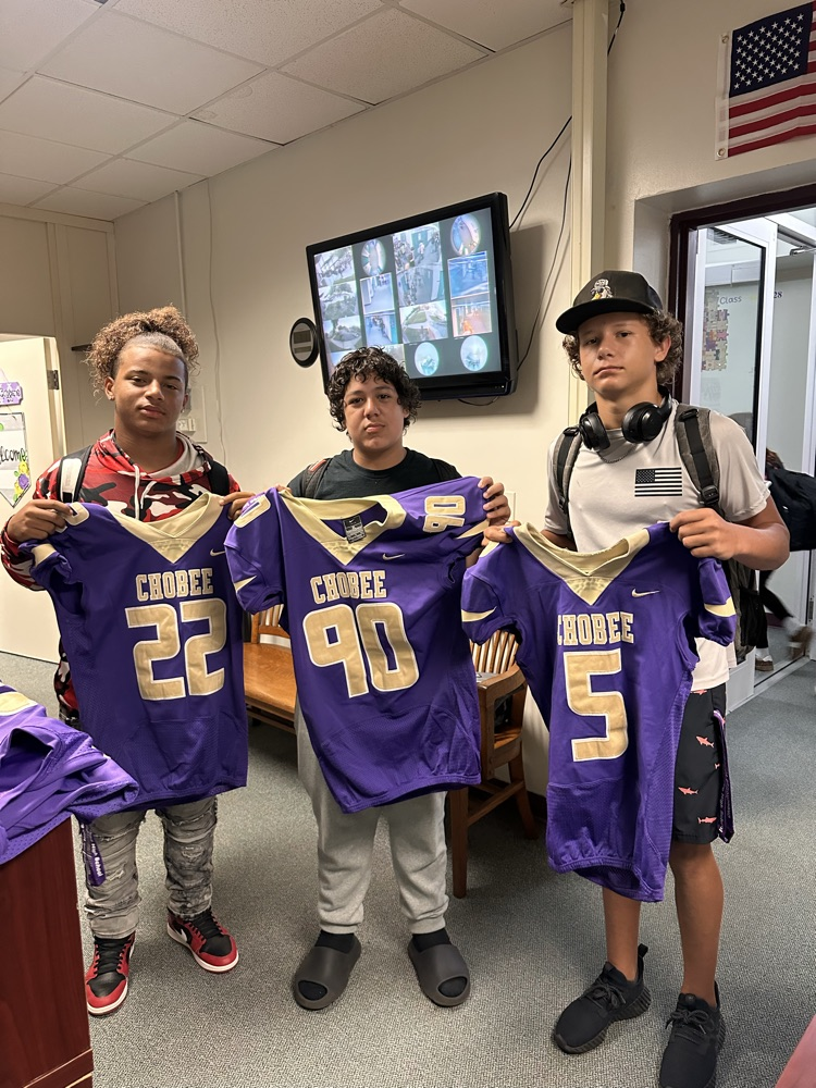 three students holding new football jersey in main office