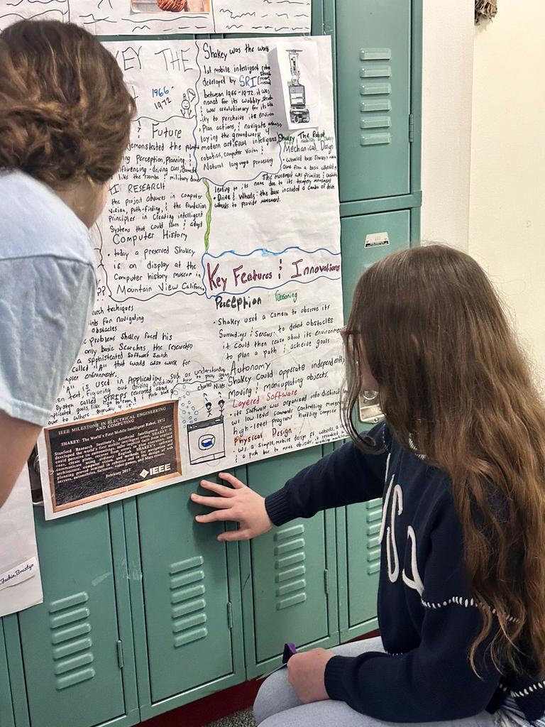 two female students presenting AI poster looking at the poster in hallway