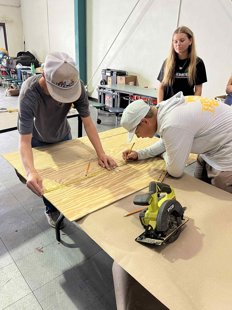 two boys measuring and writing on wood and female student supervising