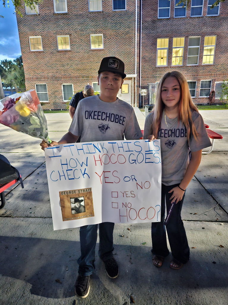 boy holding poster asking girl to homecoming dance in school courtyard