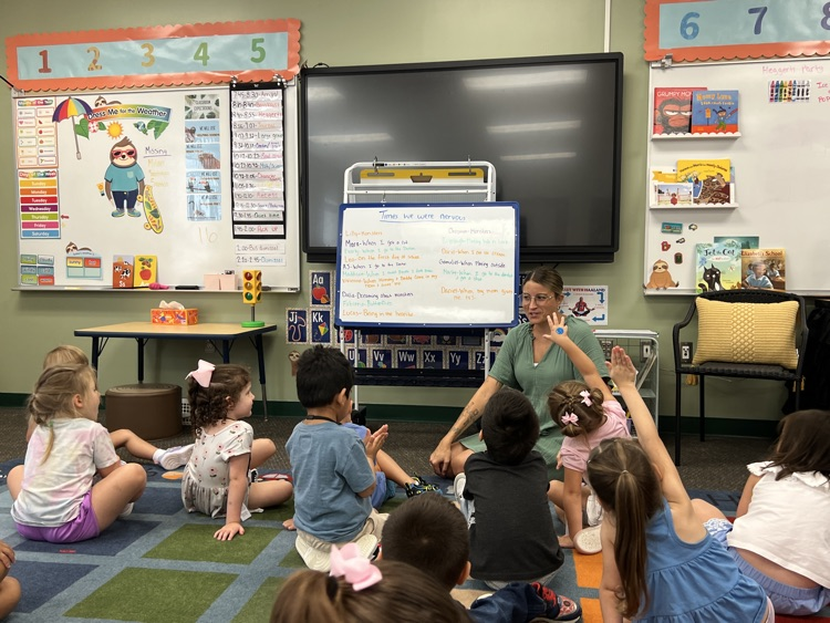 students looking at teacher sitting in rug and some raising their hand