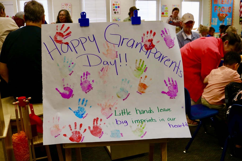 A sign with colorful handprints and text "Happy Grandparents Day!!!" in front of seated adults.