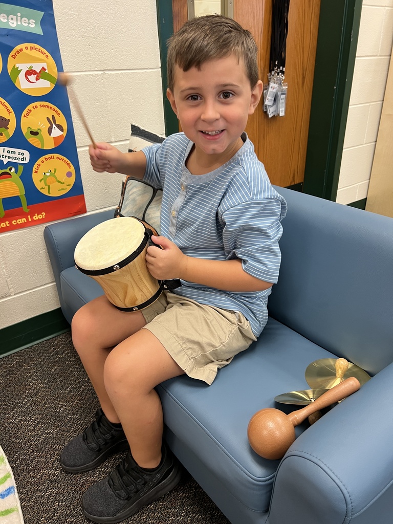 Child seated on a blue chair holding a drum and a drumstick in a classroom setting.