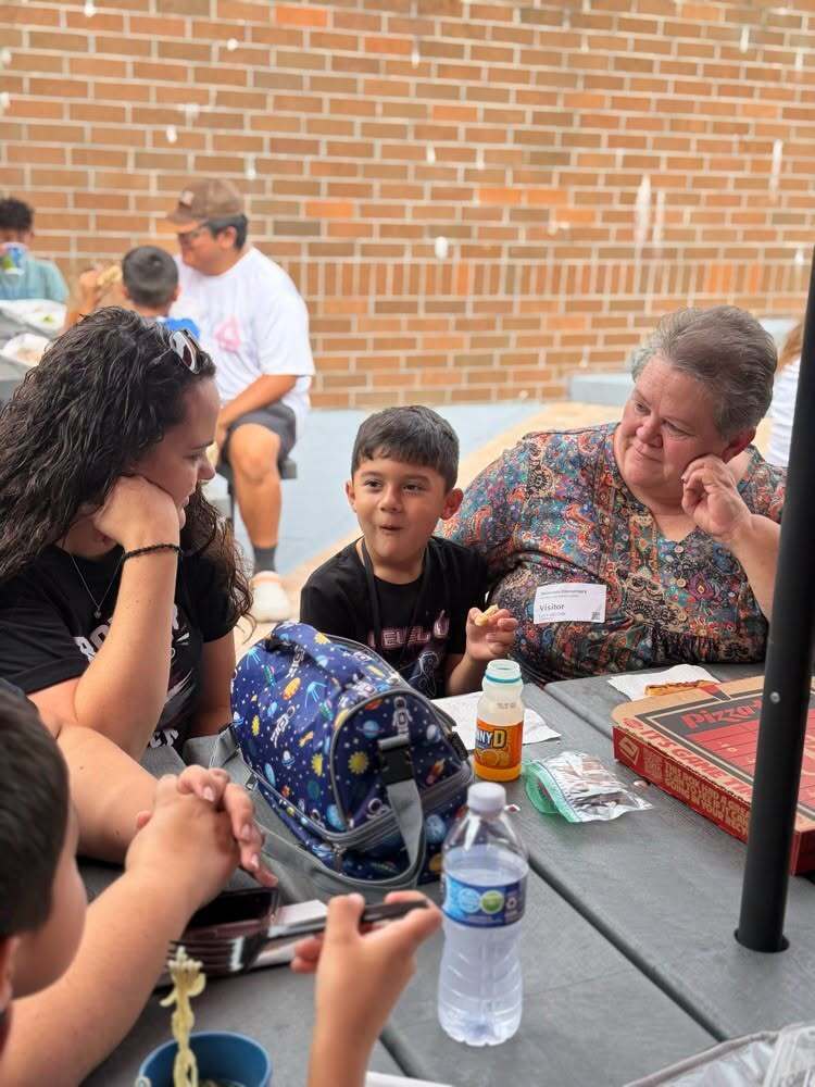 People gathered around a table, possibly outdoors. A woman talks to a child. A bag, cup, bottle, and game board are on the table.