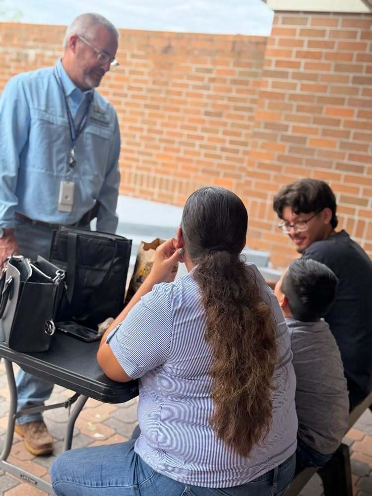 A man with glasses and a badge stands near a table. A woman with long hair listens to him. Two people sit on a bench.