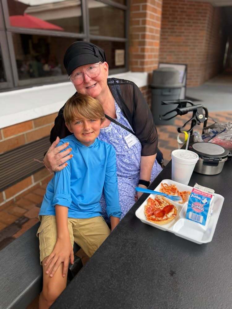 A woman and boy sit on a bench, smiling. A table holds a tray with pizza, juice, and other items.