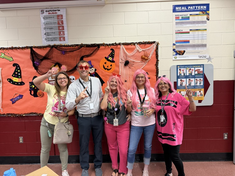 Six people standing in front of a halloween banner. Each person wearing different colored wigs and glasses.