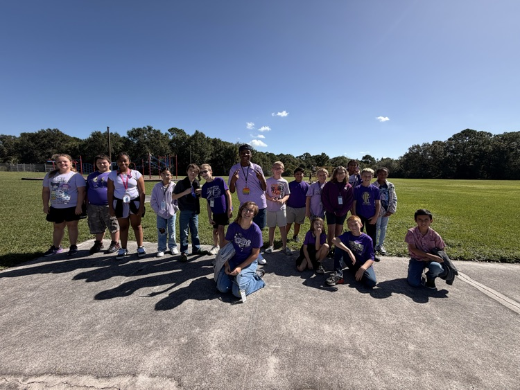 A group of students in purple shirts poses for a photo on the pavement with grass.