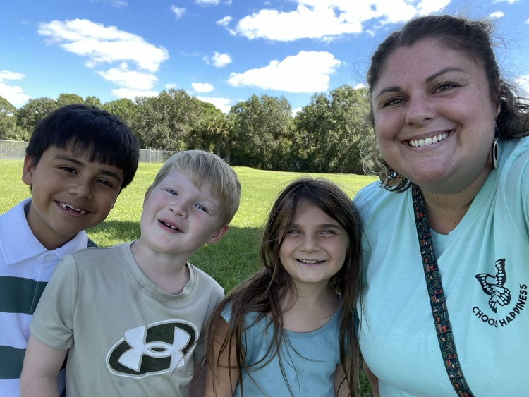 Woman smiling, standing with three children in a grassy field with trees in the background.