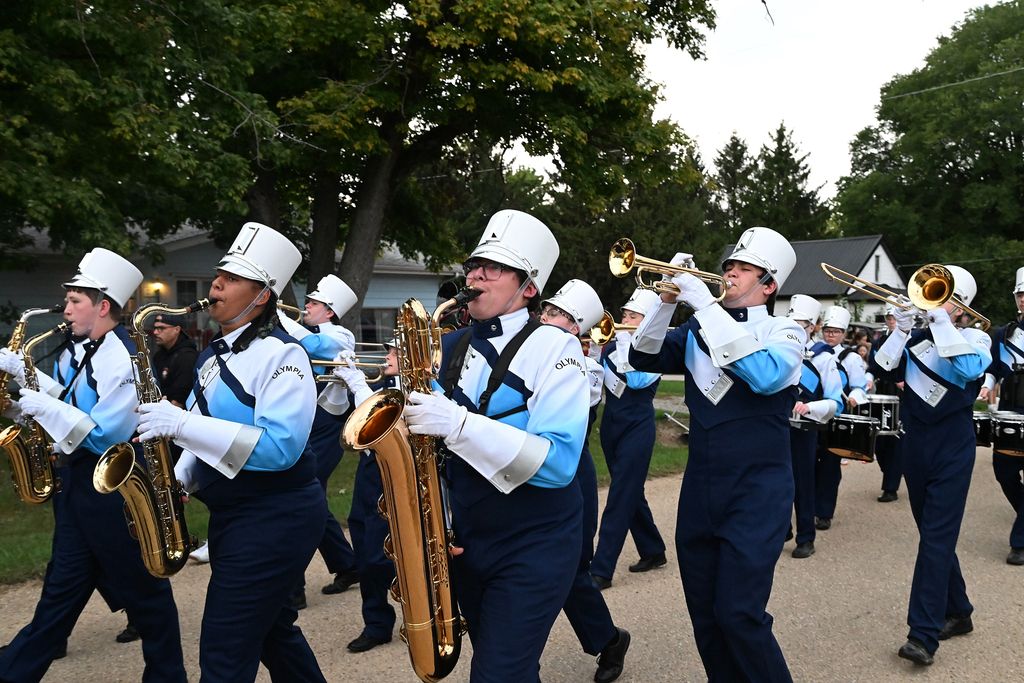 A marching band walks down the street playing instruments in blue and white uniforms