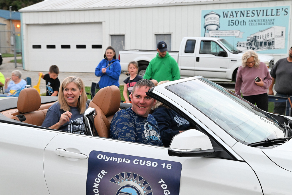 A man and women sit in a white convertible smiling at a crowd