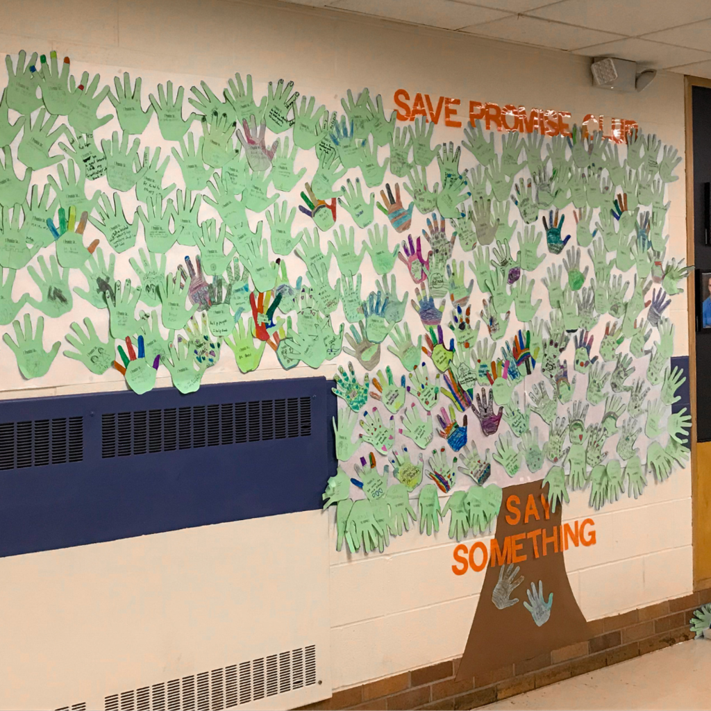 Wall covered in cutout paper hands and a sign that says "Save Promise Club" and "say something."