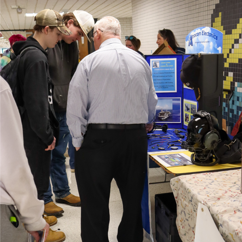 two students speaking with a man in front of a table.