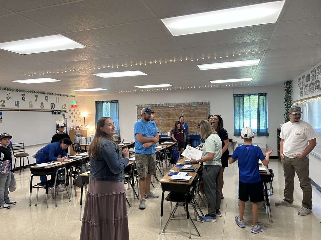 Adults and children gather among classroom desks during a school visit, talking and reviewing materials.