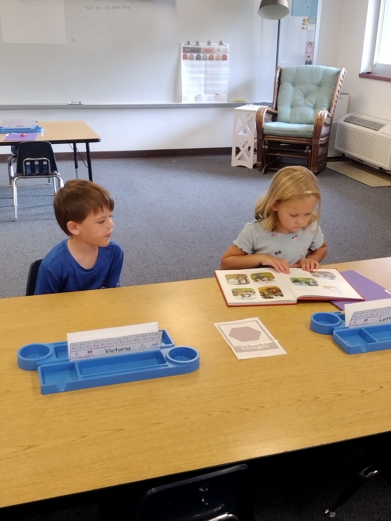 Two young students sit at a classroom table, with one looking on as the other reads an illustrated book.