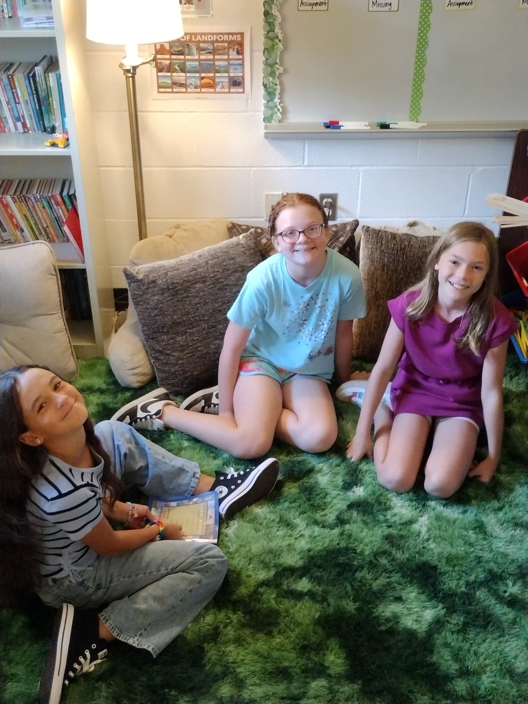 Three smiling students sit together on a soft green rug in a classroom reading area surrounded by cushions.