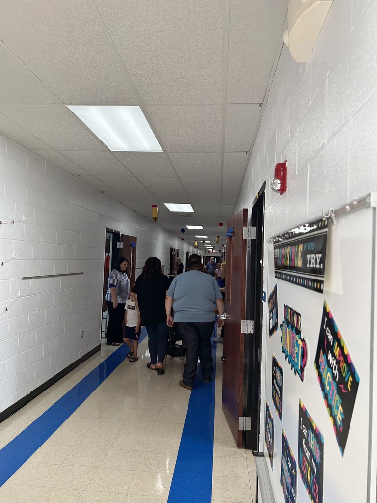 Adults and children walk through a brightly lit school hallway lined with classrooms and colorful displays.