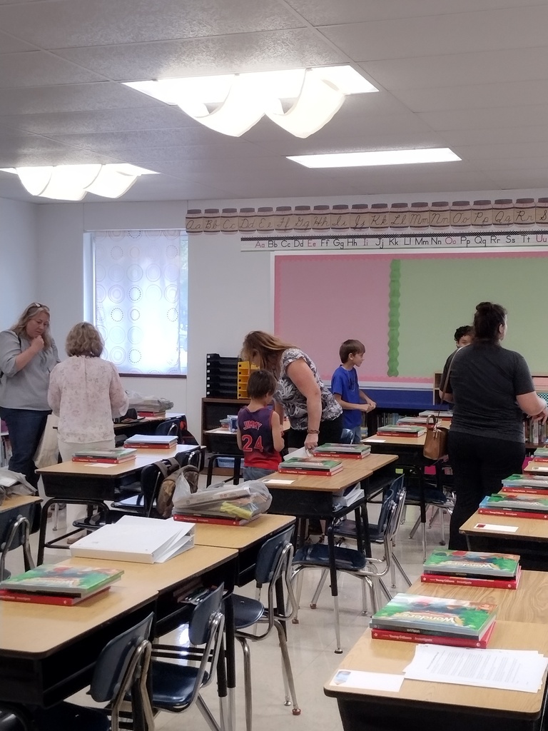 Adults and children explore a classroom where textbooks and supplies are neatly arranged on student desks.