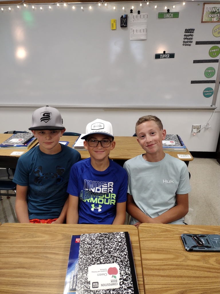Three smiling students sit together at classroom desks with notebooks and school supplies in front of them.