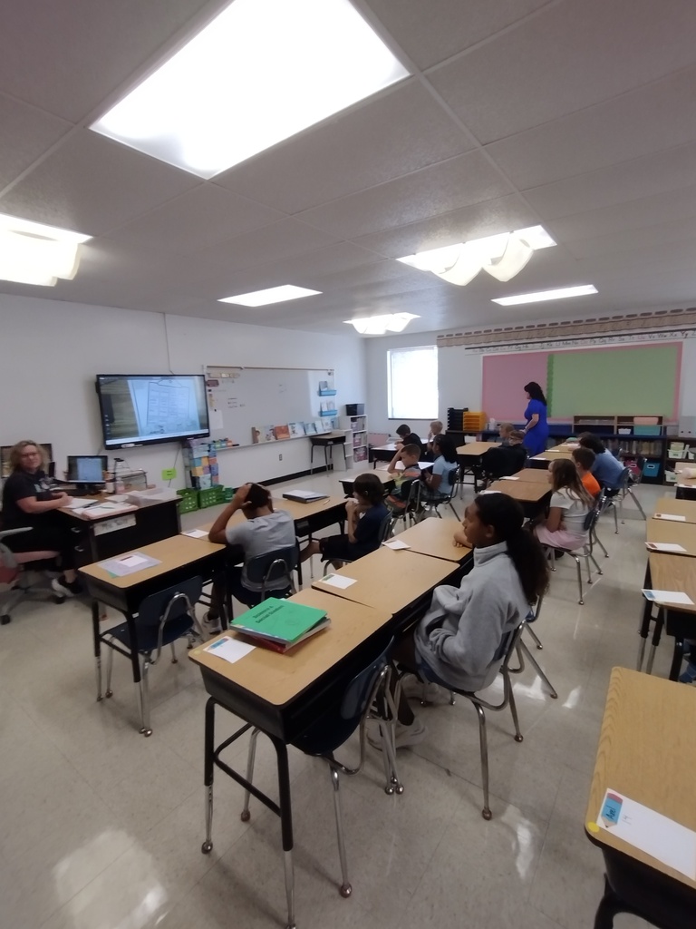 Children sitting at desks in a classroom listening to a teacher speak.