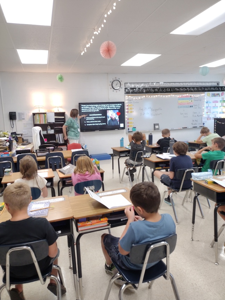 Children sitting at desks in a classroom listening to a teacher present on a screen.